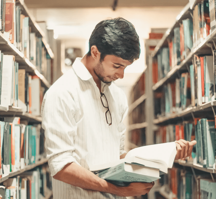Student o czarnych włosach, ubrany w białą koszulę czyta książkę, stojąc między regałami biblioteki, na których stoją książki.