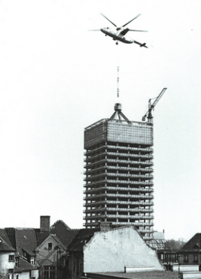 Historyczne czarno-białe zdjęcie budynku altum podczas budowy. Nad altum widnieje helikopter opuszczający fragment budynku.
