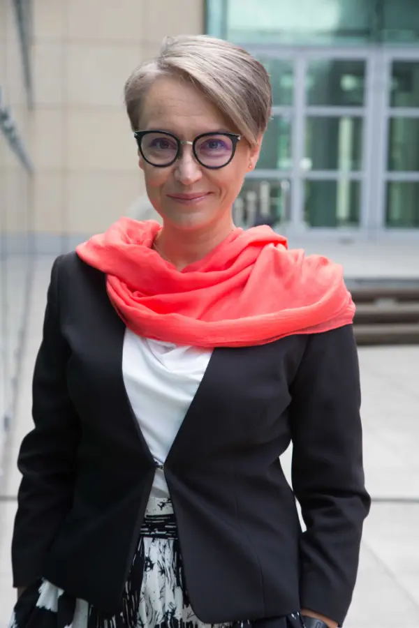 Kobieta z krótkimi, jasnymi włosami, ubrana w czarną marynarkę i biały top, z jasnoczerwonym szalikiem na szyi, stoi przed nowoczesnym budynkiem. Ma na sobie okrągłe okulary i lekki uśmiech na twarzy.