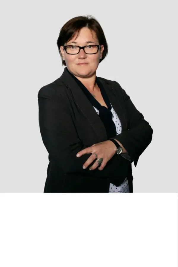 Kobieta z krótkimi, ciemnymi włosami, nosząca okulary, ubrana w czarny żakiet i białą bluzkę w kropki, stoi na jasnym tle. Jej ręce są skrzyżowane na piersi, a na twarzy ma pewny siebie wyraz.