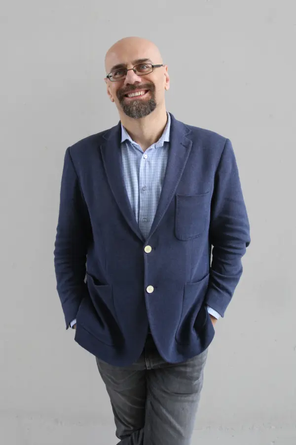 Prof. Jacek Marszałek w granatowej marynarce i niebieskiej koszuli, z okularami na nosie i uśmiechnięty, stoi na szarym tle. Ma ręce schowane w kieszeniach, sprawiając wrażenie zrelaksowanego i przyjaznego.