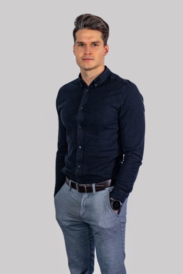 Portret Michała Kowalskiego, mężczyzny w średnim wieku, ubranej w ciemnoniebieską koszulę i szare spodnie. Michał stoi z rękami w kieszeniach, patrząc prosto w kamerę, na neutralnym, szarym tle.