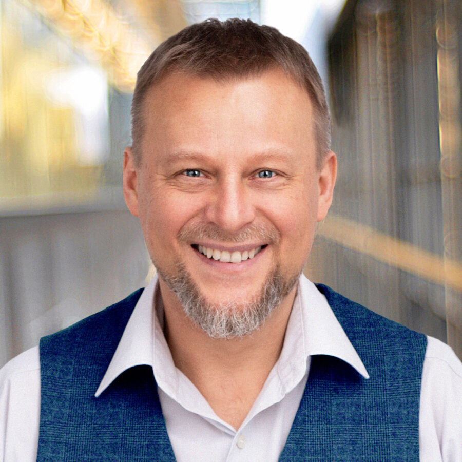 Portret mężczyzny o jasnych włosach i krótkiej brodzie, uśmiechającego się szeroko do obiektywu. Jest ubrany w białą koszulę i niebieską kamizelkę. Tło jest rozmyte, co sprawia, że mężczyzna jest głównym punktem zdjęcia.