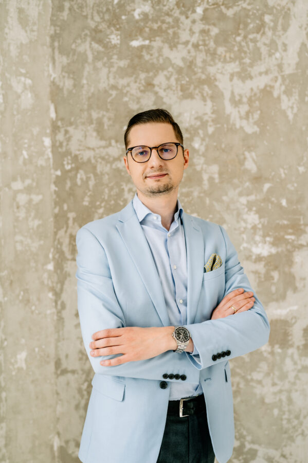 Portret Krzysztofa Góreckiego, mężczyzny w średnim wieku, ubranego w jasnoniebieską marynarkę, błękitną koszulę i ciemne spodnie. Krzysztof stoi z założonymi rękami na tle ściany o surowej, betonowej fakturze. Na jego twarzy widoczny jest delikatny uśmiech, a na nadgarstku nosi elegancki zegarek.