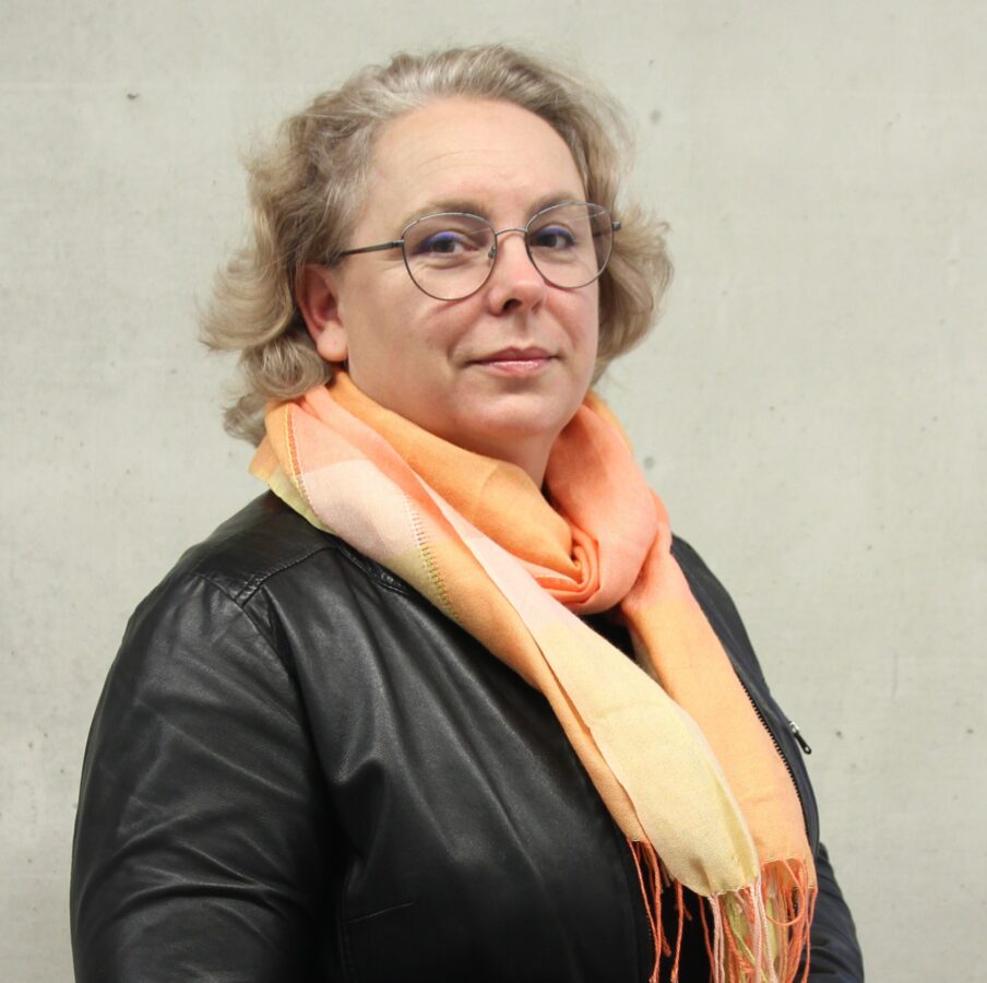 Portret kobiety w średnim wieku, noszącej okulary i krótko ścięte, siwe włosy. Ubrana jest w czarną, skórzaną kurtkę i jasnopomarańczowy szalik. Kobieta spogląda bezpośrednio w obiektyw, a za nią widać neutralne tło w kolorze jasnoszarym.