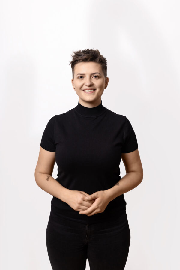 Uśmiechnięta kobieta z krótkimi włosami, ubrana w czarny, przylegający sweter z krótkim rękawem. Stoi na białym tle, trzymając ręce splecione przed sobą. Ma subtelny makijaż i kolczyki. Na jej lewym przedramieniu widoczny jest tatuaż.