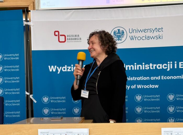 Zdjęcie przedstawia kobietę stojącą przy mównicy podczas konferencji na Uniwersytecie Wrocławskim. Kobieta trzyma żółty mikrofon i uśmiecha się, ubrana jest w ciemne ubranie i ma na szyi identyfikator na niebieskiej smyczy. W tle widoczne są banery Uniwersytetu Wrocławskiego z logo uczelni oraz napisem "Wydział Prawa, Administracji i Ekonomii". Na mównicy znajdują się dwa plakaty promujące Międzynarodową Konferencję Naukową "Zdrowie i Styl Życia Wrocław 2024". W górnej części zdjęcia widać fragment ekranu projekcyjnego.