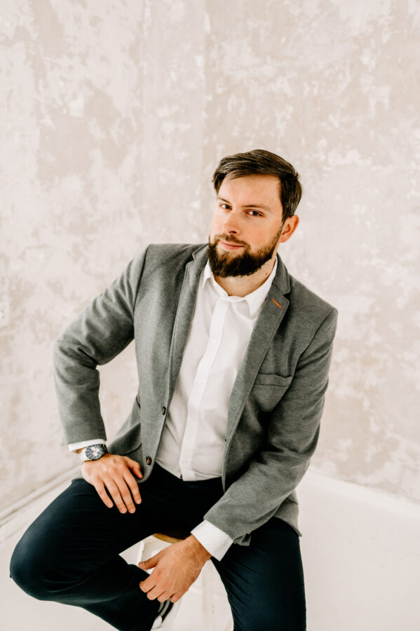Portret mężczyzny na tle jasnej, lekko teksturowanej ściany. Mężczyzna ma krótkie, ciemne włosy i brodę. Nosi szary garnitur, białą koszulę i ciemne spodnie. Siedzi na stołku, opierając jedną rękę na kolanie, a drugą na biodrze. Jego wyraz twarzy jest spokojny, a wzrok skierowany lekko w bok. Na ręce ma zegarek.