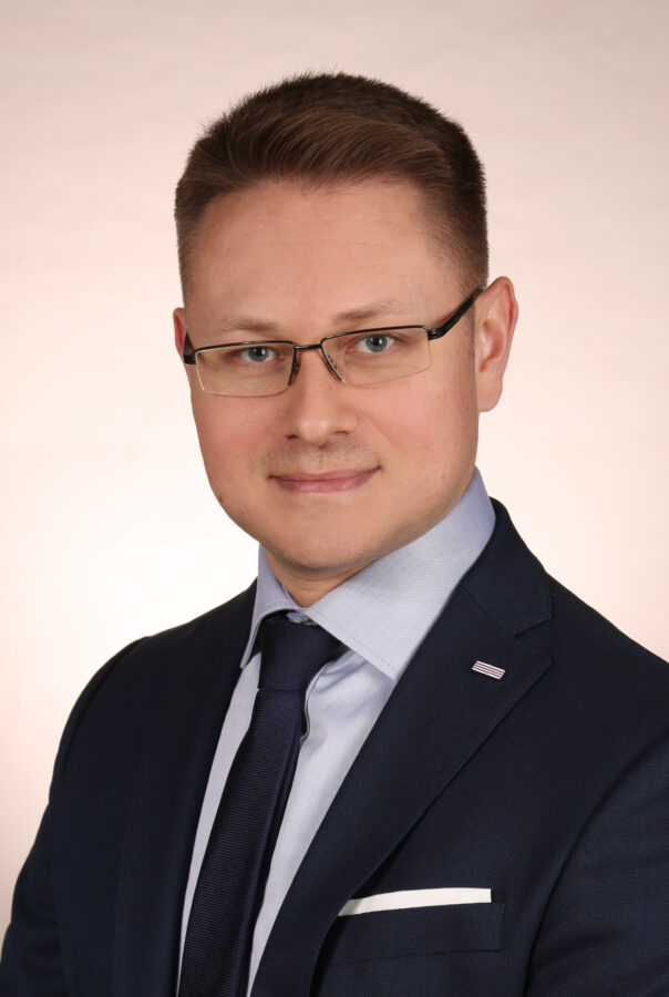 Marcin Gurtatowski, mężczyzna w średnim wieku, noszący okulary, ubrany w ciemny garnitur z jasnoniebieską koszulą i granatowym krawatem. Tło zdjęcia jest jednolite, jasne, co podkreśla profesjonalny charakter portretu. Marcin Gurtatowski patrzy prosto w obiektyw, uśmiechając się lekko.