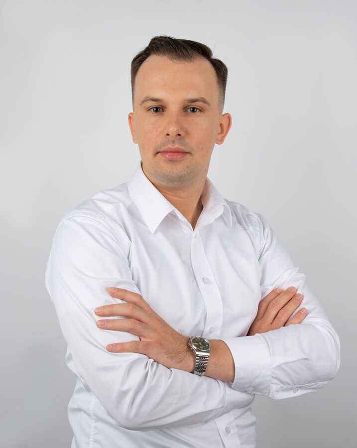 Mężczyzna z krótkimi, ciemnymi włosami, ubrany w białą koszulę, stoi z założonymi rękoma na piersi. Na jego lewym nadgarstku znajduje się zegarek. Tło jest jednolite, jasnoszare, co nadaje zdjęciu profesjonalny charakter. Mężczyzna patrzy prosto w kamerę, emanując pewnością siebie i spokojem.