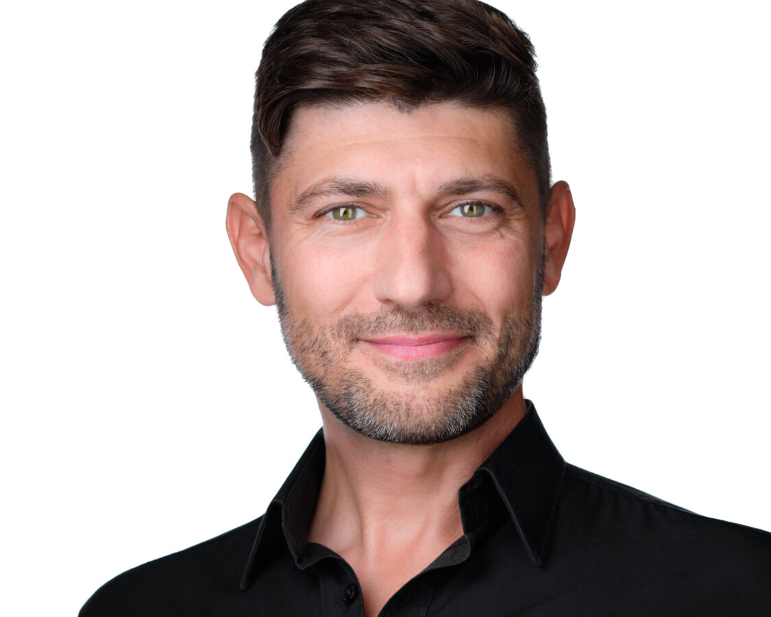 Portret mężczyzny na białym tle. Ma krótkie, ciemne włosy i lekki zarost. Nosi czarną koszulę z kołnierzykiem i uśmiecha się delikatnie, patrząc prosto w kamerę.