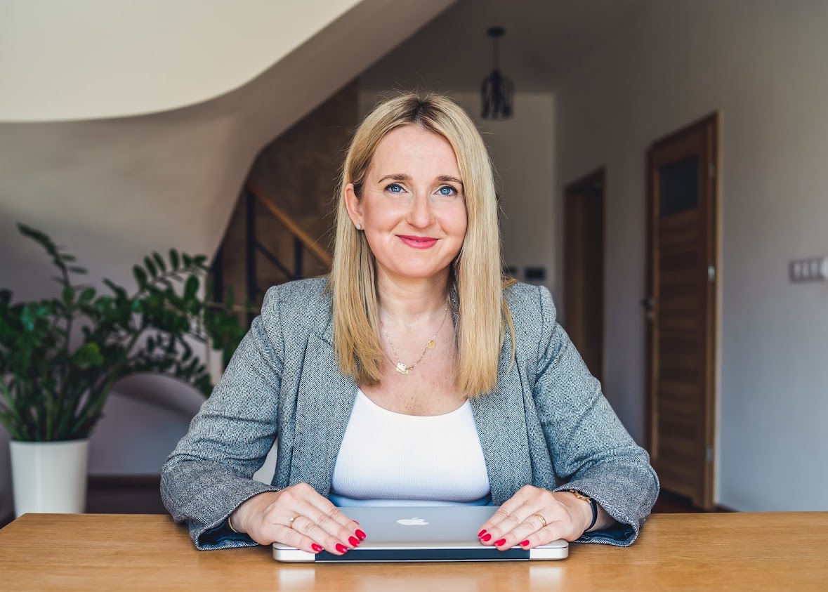 Portret kobiety siedzącej przy stole z laptopem. Ma blond włosy i uśmiech na twarzy, nosi szary żakiet i białą bluzkę. W tle widać wnętrze domu z rośliną w doniczce i drewnianymi drzwiami.