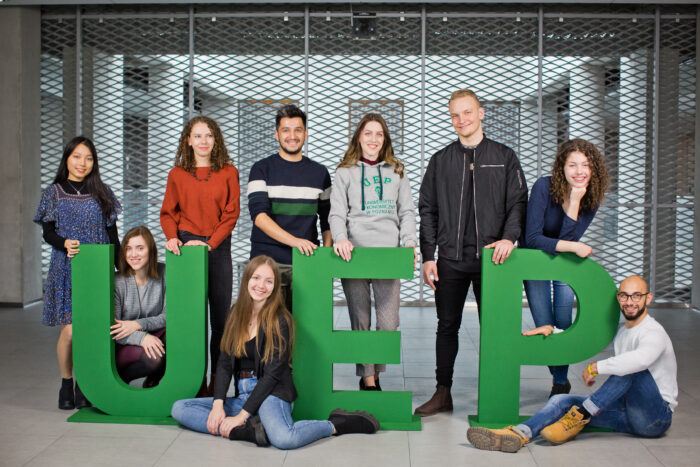 Na zdjęciu znajduje się grupa studentów Uniwersytetu Ekonomicznego w Poznaniu, którzy pozują z dużymi, zielonymi literami układającymi się w skrót "UEP". Każdy z nich przyjmuje swobodną, radosną pozę, co tworzy przyjazną i pozytywną atmosferę. Uczniowie są różnorodni, co podkreśla międzynarodowy charakter uczelni. Tło zdjęcia to nowoczesne wnętrze uniwersytetu z metalową siatką, co dodaje zdjęciu industrialnego charakteru. Wszyscy studenci wyglądają na zadowolonych i zaangażowanych, co może sugerować ich dumę z bycia częścią społeczności UEP.