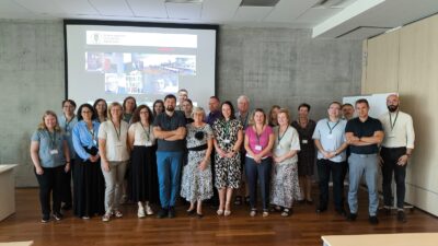 Zdjęcie przedstawia grupę kilkunastu osób stojących w sali konferencyjnej. W tle widoczny jest ekran z wyświetlonymi obrazami z wideokonferencji.