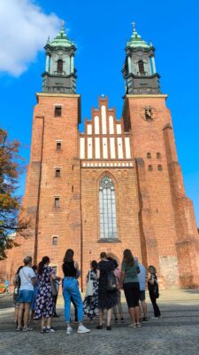 Zdjęcie przedstawia grupę osób stojących przed monumentalną fasadą katedry w Poznaniu z charakterystycznymi dwoma wieżami i dużym gotyckim oknem w centralnej części.