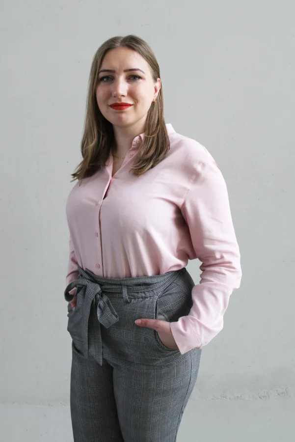 Portret kobiety w różowej koszuli i szarych spodniach w kratę. Kobieta ma długie, jasne włosy i czerwone usta. Tło jest jasne, jednolite.