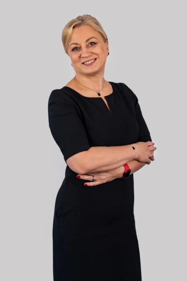 Uśmiechnięta kobieta z blond włosami spiętymi do tyłu, ubrana w ciemnogranatową sukienkę. Stoi z założonymi rękami na tle jasnoszarej ściany, nosi biżuterię i zegarek.