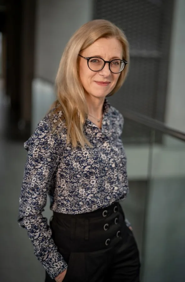 Kobieta z długimi blond włosami i okularami, ubrana w ciemną koszulę w jasne kwiaty. Uśmiecha się delikatnie, stoi przy szklanej balustradzie wewnątrz nowoczesnego budynku.