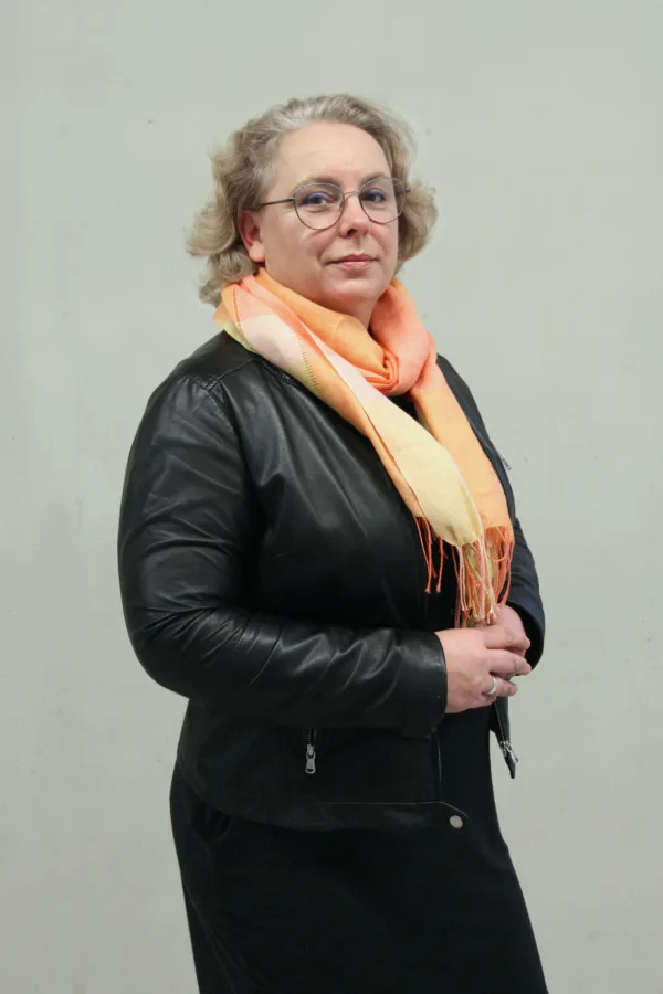 Kobieta w okularach, ubrana w czarną skórzaną kurtkę i pomarańczowo-żółty szalik, stoi na tle jasnej ściany. Ma krótkie, falowane włosy i spokojny wyraz twarzy.