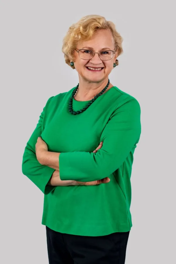 Mężczyzna z brodą i okularami ubrany w granatową marynarkę, zielony krawat i brązowe spodnie, stoi na tle jasnej ściany. Uśmiecha się lekko i ma dłonie oparte na biodrach.