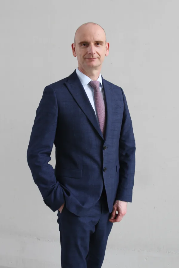 Mężczyzna w garniturze w kratę, jasnoniebieskiej koszuli i jasnofioletowym krawacie stoi na tle jasnej ściany. Ma ogoloną głowę i delikatny uśmiech.