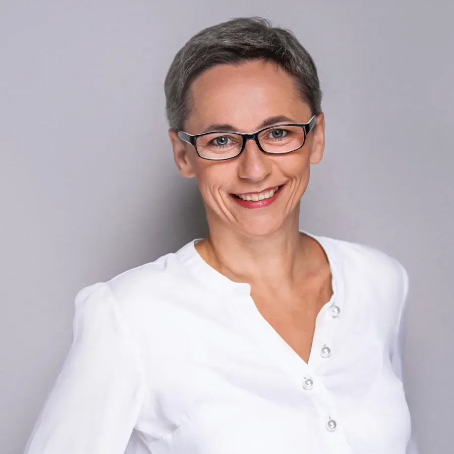 Kobieta z krótkimi siwymi włosami i okularami uśmiecha się do aparatu. Ubrana w białą koszulę z guzikami, stoi na tle jednolitej szarej ściany. Zdjęcie portretowe, profesjonalne, o jasnym i przyjaznym charakterze.