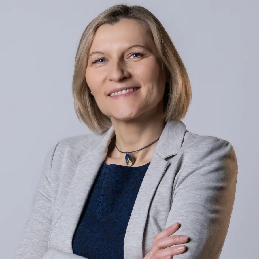Kobieta w jasnej marynarce i granatowej bluzce pozuje na neutralnym tle. Ma blond włosy do ramion, delikatny makijaż, uśmiech i założone ręce. Nosi naszyjnik z kolorowym wisiorkiem.
