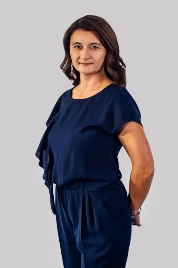 Kobieta w granatowym stroju stoi na tle jasnoszarego tła. Ma ciemne, falowane włosy i delikatny uśmiech.