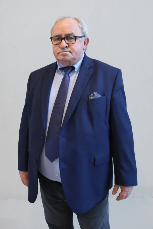 Mężczyzna w średnim wieku w granatowej marynarce i krawacie stoi na tle jasnej ściany. Ma okulary i poważny wyraz twarzy.