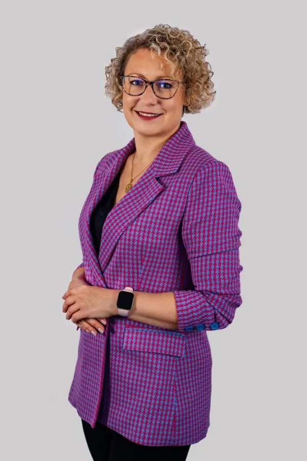 Uśmiechnięta kobieta z kręconymi blond włosami i okularami, ubrana w różowo-niebieską marynarkę w jodełkę. Ma na ręku smartwatch i złoty naszyjnik. Stoi na tle jasnoszarej ściany.
