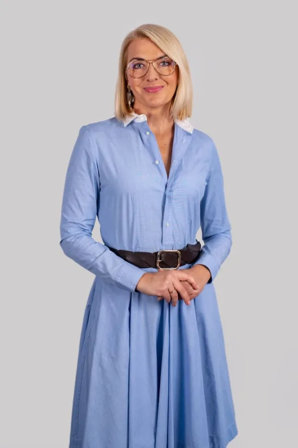 Kobieta o jasnej karnacji i blond włosach do ramion, z przedziałkiem po lewej stronie, uśmiecha się i patrzy wprost. Ma na sobie jasnoniebieską sukienkę koszulową z długimi rękawami i białym kołnierzykiem, przewiązaną w talii szerokim, ciemnobrązowym paskiem ze złotą klamrą. Nosi okulary w metalowych oprawkach typu aviator oraz długie, wiszące kolczyki w złotym kolorze. Jej dłonie są splecione przed nią. Tło jest jednolite, w jasnoszarym kolorze.