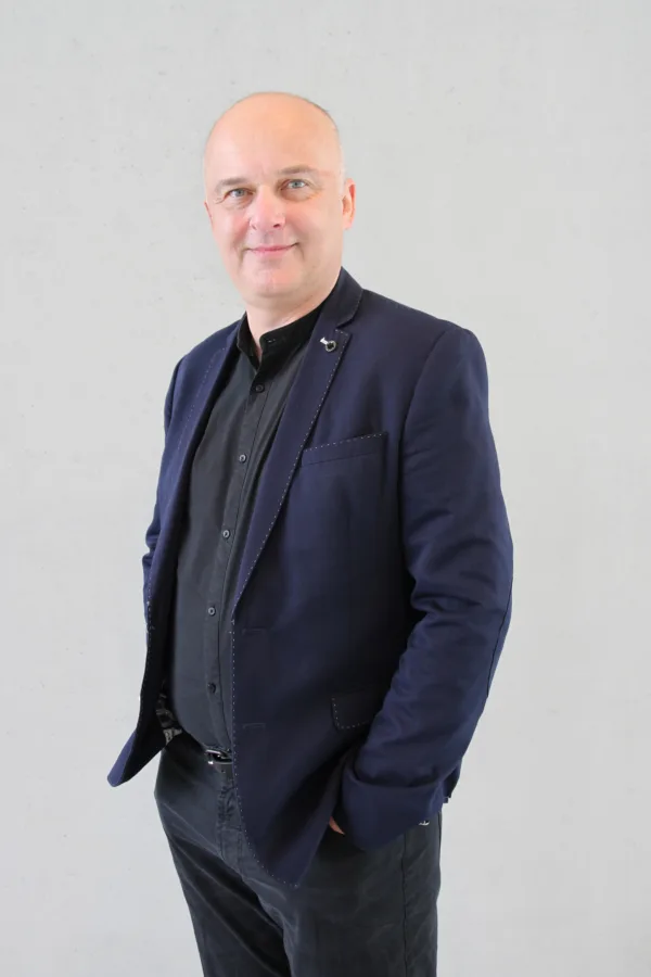 Uśmiechnięty mężczyzna z ogoloną głową, ubrany w ciemną koszulę i granatową marynarkę ze zdobionymi przeszyciami. Stoi na tle jasnej ściany z rękami w kieszeniach.