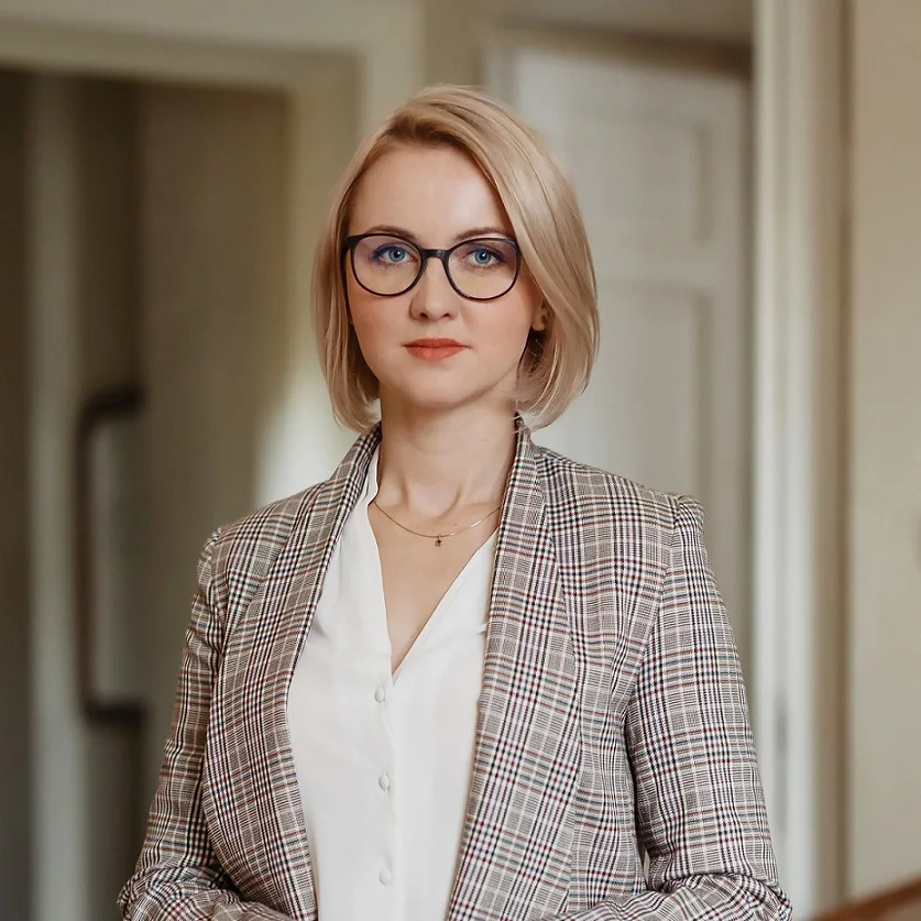 Portret kobiety w okularach, z jasnymi włosami do ramion, ubranej w białą bluzkę i marynarkę w kratę. Kobieta patrzy wprost, tło jest jasne, rozmyte.
