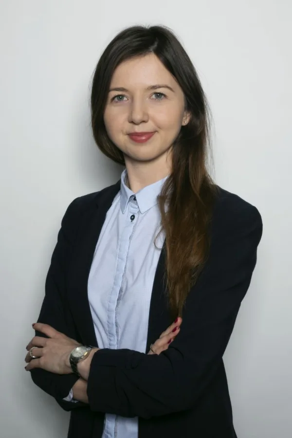 Młoda kobieta z długimi, brązowymi włosami, ubrana w jasnoniebieską koszulę i ciemną marynarkę. Ma założone ręce, lekki uśmiech i stoi na tle jasnej ściany.