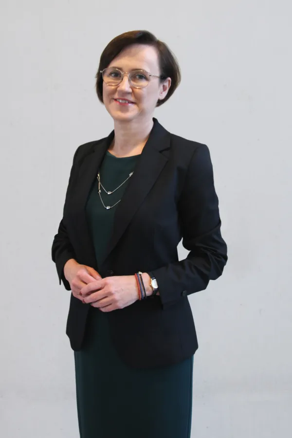 Uśmiechnięta kobieta w okularach, ubrana w ciemną marynarkę i zieloną sukienkę. Nosi delikatny naszyjnik, zegarek i kolorowe bransoletki. Stoi na tle jasnej ściany.