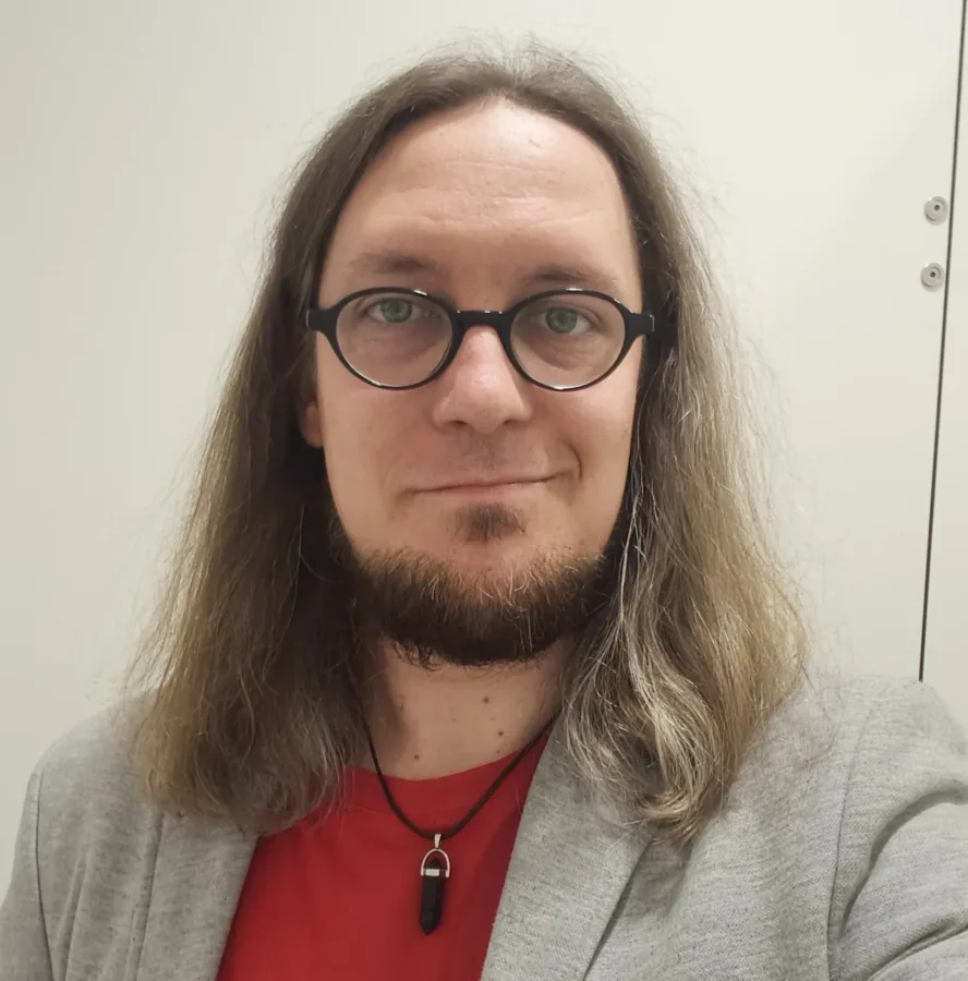 Portret mężczyzny z długimi, jasnobrązowymi włosami, brodą i wąsami. Nosi czarne, okrągłe okulary, czerwoną koszulkę i szarą marynarkę. Na szyi ma czarny sznurek z wisiorkiem w kształcie czarnego kamienia. Mężczyzna lekko się uśmiecha i patrzy wprost. Tło jest jasne, jednolite.