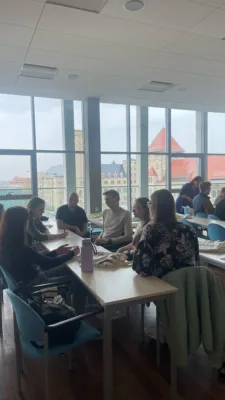 W jasnej sali z dużymi oknami Uniwersytetu Ekonomicznego w Poznaniu studenci siedzą przy stolikach, pracując w grupach. Dyskutują, piszą i przeglądają materiały. Na stołach leżą notatki i butelki z wodą. Za oknami widoczny jest miejski krajobraz Poznania, w tym budynki z czerwonymi dachami.