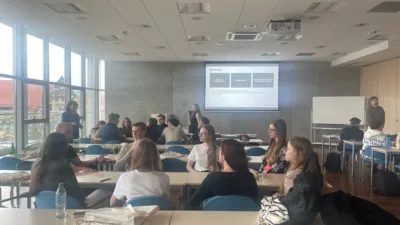 W sali wykładowej Uniwersytetu Ekonomicznego w Poznaniu trwa interaktywne zajęcie. Na ekranie projekcyjnym wyświetlony jest slajd z tytułem "Wnioski" i schematem. Studenci siedzą przy stolikach w grupach, dyskutując i pracując. W sali panuje ożywiona atmosfera. Widoczne są duże okna z widokiem na Poznań oraz biała tablica.
