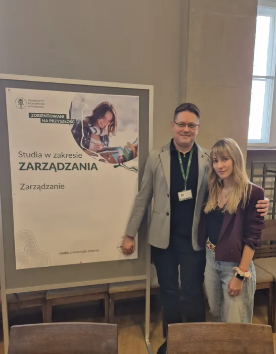 W Poznaniu mężczyzna w okularach i szarej marynarce oraz młoda kobieta z blond włosami w bordowej marynarce stoją obok plakatu promującego studia na kierunku ZARZĄDZANIE na Uniwersytecie Ekonomicznym w Poznaniu. Plakat ma jasne tło i zawiera zdjęcie uśmiechniętej studentki pracującej przy biurku. W górnej części plakatu widoczne jest logo UEP oraz hasło "ZORIENTOWANI NA PRZYSZŁOŚĆ". Pod tytułem "Studia w zakresie ZARZĄDZANIA" znajduje się dodatkowy napis "Zarządzanie". Para uśmiecha się do kamery.
