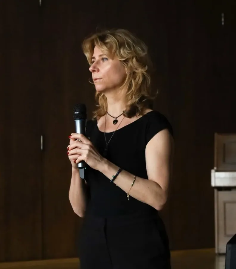 Kobieta o jasnych, kręconych włosach trzyma mikrofon w dłoniach i patrzy w lewą stronę. Ubrana jest w czarną sukienkę z krótkim rękawem i ma na sobie kilka cienkich naszyjników oraz bransoletki na obu nadgarstkach. Jej paznokcie są pomalowane na czerwono. Tło za nią jest ciemne, w odcieniach brązu.