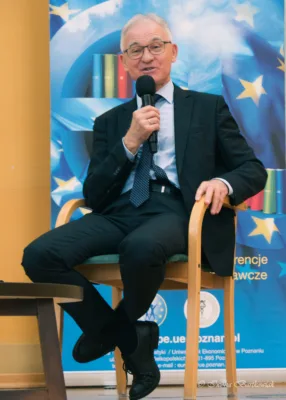 Starszy mężczyzna z siwymi włosami i okularach siedzi na drewnianym krześle i mówi do mikrofonu, trzymając go w prawej dłoni. Ubrany jest w ciemny garnitur, jasną koszulę i wzorzysty krawat. Jego nogi są skrzyżowane. W tle widoczny jest baner z motywami Unii Europejskiej (gwiazdki na niebieskim tle) oraz napisy w języku polskim. Mężczyzna patrzy w stronę słuchaczy i wydaje się przemawiać.