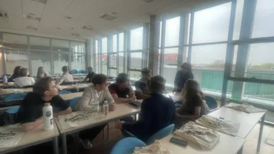 W jasnej sali z dużymi oknami Uniwersytetu Ekonomicznego w Poznaniu studenci pracują w grupach przy stolikach. Na stołach leżą notatki, długopisy i butelki wody. Niektórzy studenci rozmawiają, inni piszą lub przeglądają materiały. Na niektórych stolikach widoczne są torby z logo "Carlsberg Group".