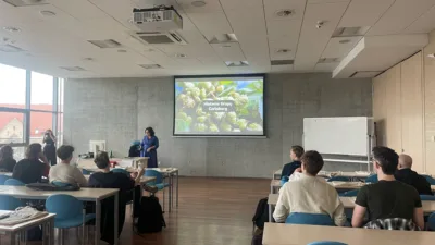 W jasnej sali wykładowej Uniwersytetu Ekonomicznego w Poznaniu trwa prezentacja. Na ekranie projekcyjnym wyświetlony jest slajd zatytułowany "Historia Grupy Carlsberg" z grafiką przedstawiającą chmiel. Kobieta w niebieskiej sukience stoi obok ekranu i gestykuluje, prowadząc wykład dla siedzących przy stolikach studentów. W sali znajduje się również biała tablica.