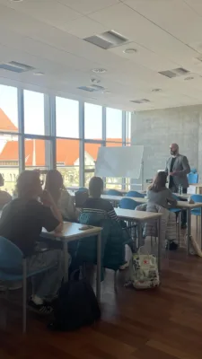 W jasnej sali wykładowej, prawdopodobnie na Uniwersytecie Ekonomicznym w Poznaniu, mężczyzna w szarym garniturze prowadzi zajęcia. Stoi przy biurku i mówi do grupy studentów siedzących przy oddzielnych stolikach. Studenci słuchają, niektórzy patrzą na niego, inni na swoje notatki lub telefony. Za mężczyzną widoczna jest biała tablica. Przez duże okna wpada naturalne światło, oświetlając salę i ukazując budynki za oknem.
