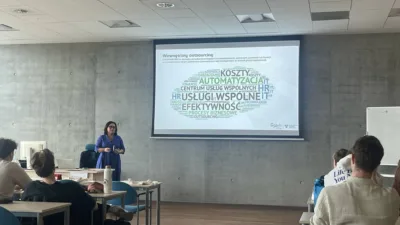 W sali wykładowej Uniwersytetu Ekonomicznego w Poznaniu trwa prezentacja na temat "Wewnętrzny outsourcing". Na ekranie projekcyjnym widoczna jest chmura wyrazów związanych z tym tematem, takich jak "KOSZTY", "AUTOMATYZACJA", "CENTRUM USŁUG WSPÓLNYCH HR", "EFEKTYWNOŚĆ". Prowadząca w niebieskiej sukience stoi obok ekranu i omawia slajd dla siedzących studentów.