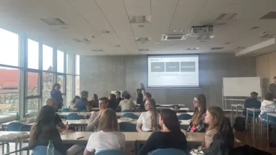 W sali wykładowej Uniwersytetu Ekonomicznego w Poznaniu trwa interaktywne zajęcie lub warsztaty. Na ekranie projekcyjnym wyświetlony jest slajd z tytułem "Wnioski" oraz schemat blokowy. Studenci siedzą przy stolikach w grupach, dyskutując i pracując nad zadaniami. W sali panuje ożywiona atmosfera. Widoczne są duże okna wpuszczające naturalne światło oraz biała tablica.