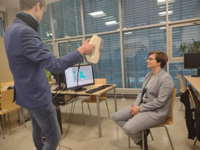 Mężczyzna trzyma ręczny skaner 3D skierowany w stronę siedzącej kobiety w jasnym garniturze. Na monitorze komputera widoczny jest tworzony w czasie rzeczywistym model 3D jej sylwetki. Sytuacja odbywa się w sali laboratoryjnej z biurkami i sprzętem komputerowym.
