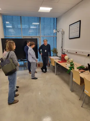 Pięć osób stoi w sali laboratoryjnej i rozmawia przy stanowisku z urządzeniami technicznymi. Na stole znajdują się modele edukacyjne, w tym dwa humanoidalne roboty oraz zielona maskotka, obok leży sprzęt elektroniczny. W tle duże okno z roletą, monitor na statywie oraz tablica z wykresem na ścianie.