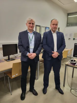 Dwóch mężczyzn w granatowych garniturach stoi obok siebie w sali komputerowej. Za nimi biurka z monitorami, na ścianie oprawione certyfikaty. Obaj mają ręce złożone przed sobą i patrzą w stronę aparatu.