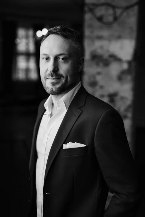 Portret w czerni i bieli przedstawiający mężczyznę w eleganckim garniturze i białej koszuli. Ujęcie półprofilowe, z miękkim światłem i płytką głębią ostrości, podkreśla spokojną pewność siebie i profesjonalizm.